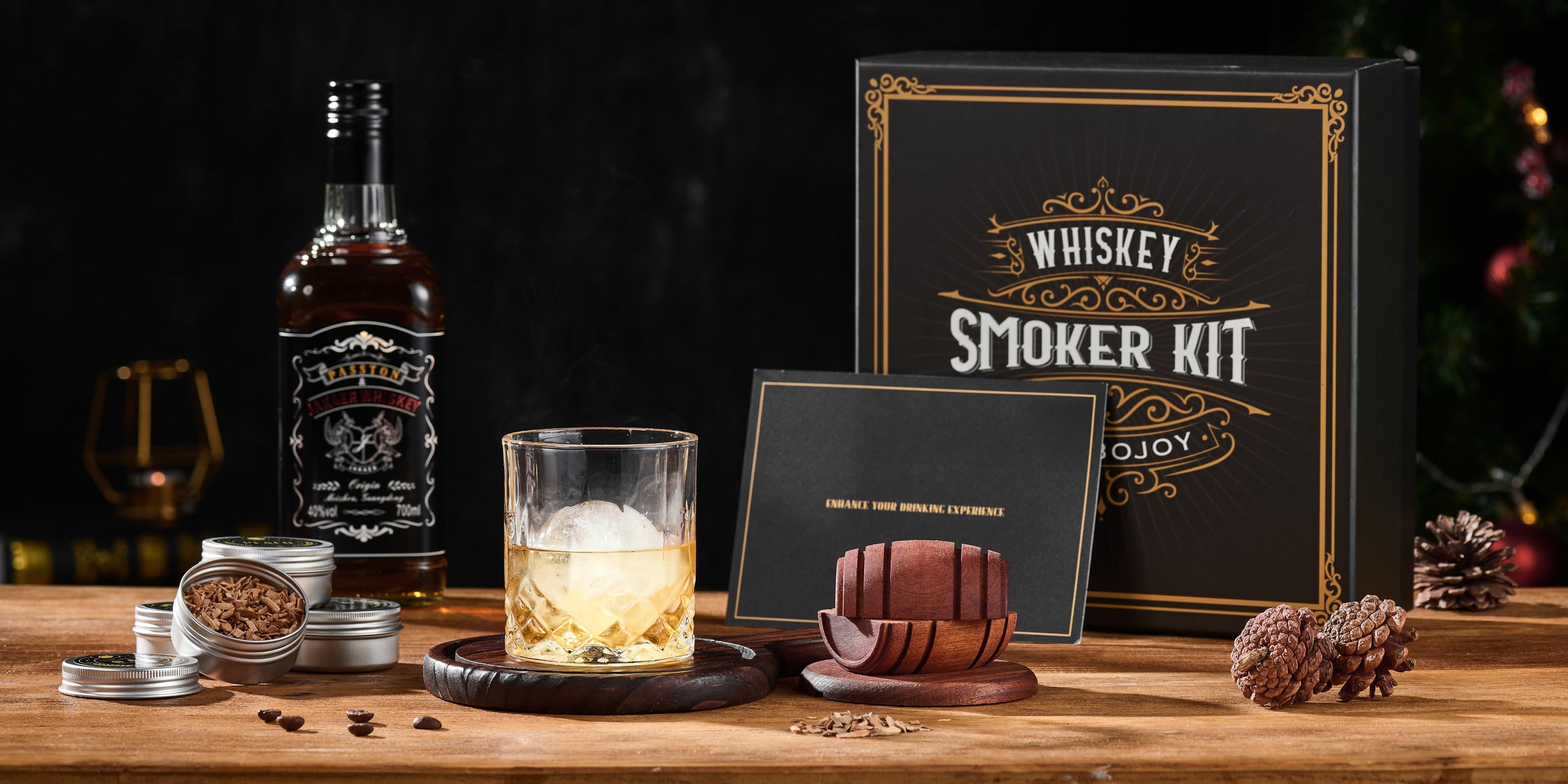 COMBOJOY barrel smoker, whiskey glass, wood chips and black gold gift box on a warmly lit wooden table
